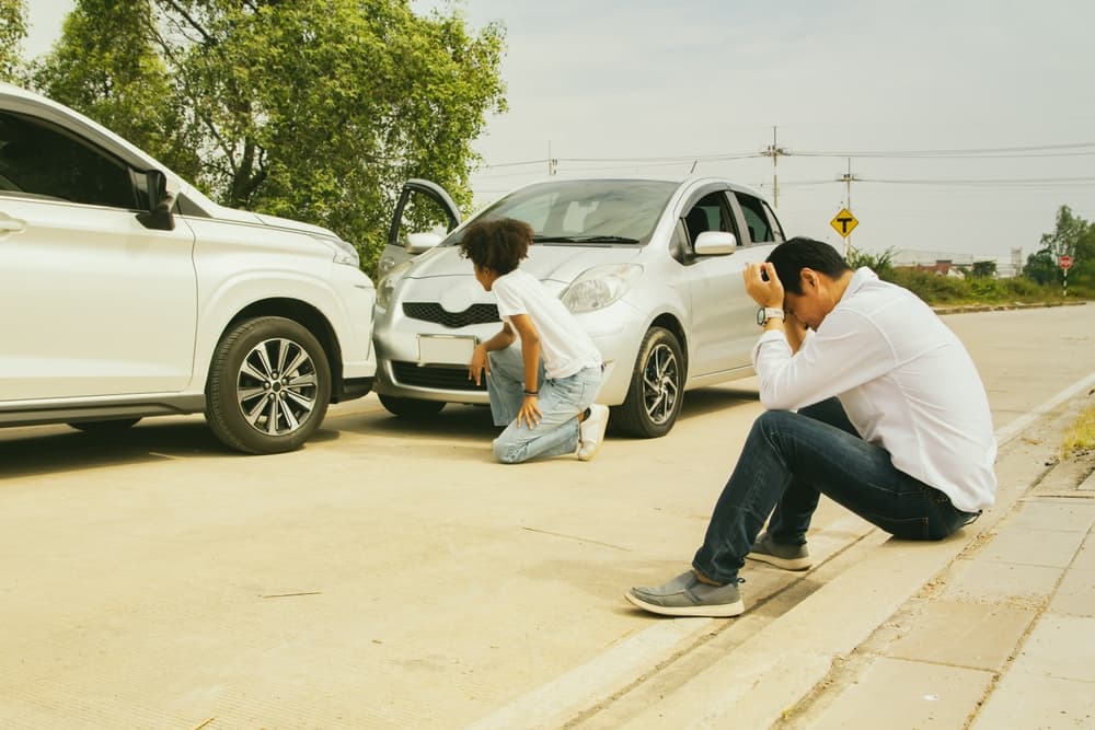 An Asian man and an African American woman, both drivers, sit stressed after a serious accident. They await insurance assistance due to illegal traffic actions that caused damage to the front bumper.