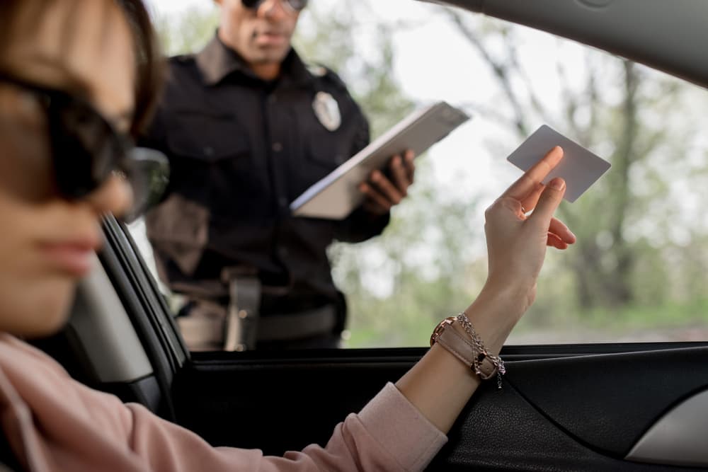 A woman wearing sunglasses sits in her car and hands her driver's license to a police officer.






